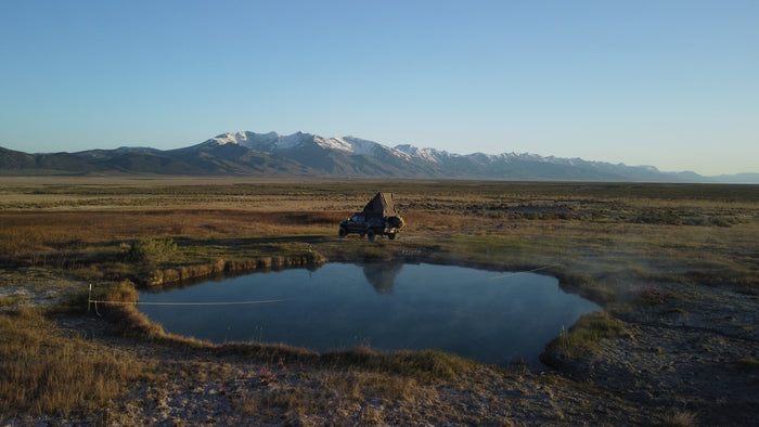 RV rental: A truck with a pop-up tent camps by a tranquil hot spring, reflecting snowy mountains and vast plains.
