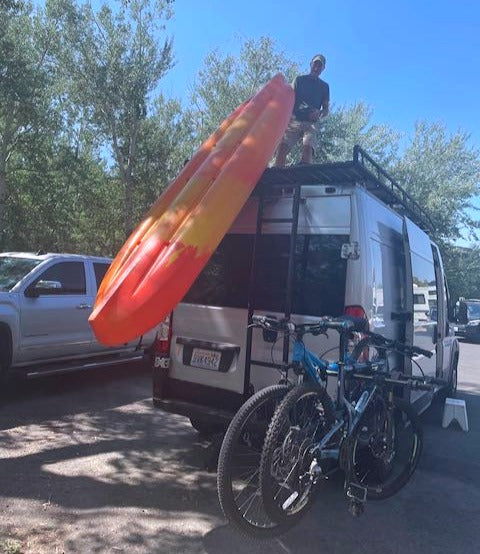 RV rental van adventure setup: A man on the roof handles a kayak, with two bikes mounted on the rear rack.