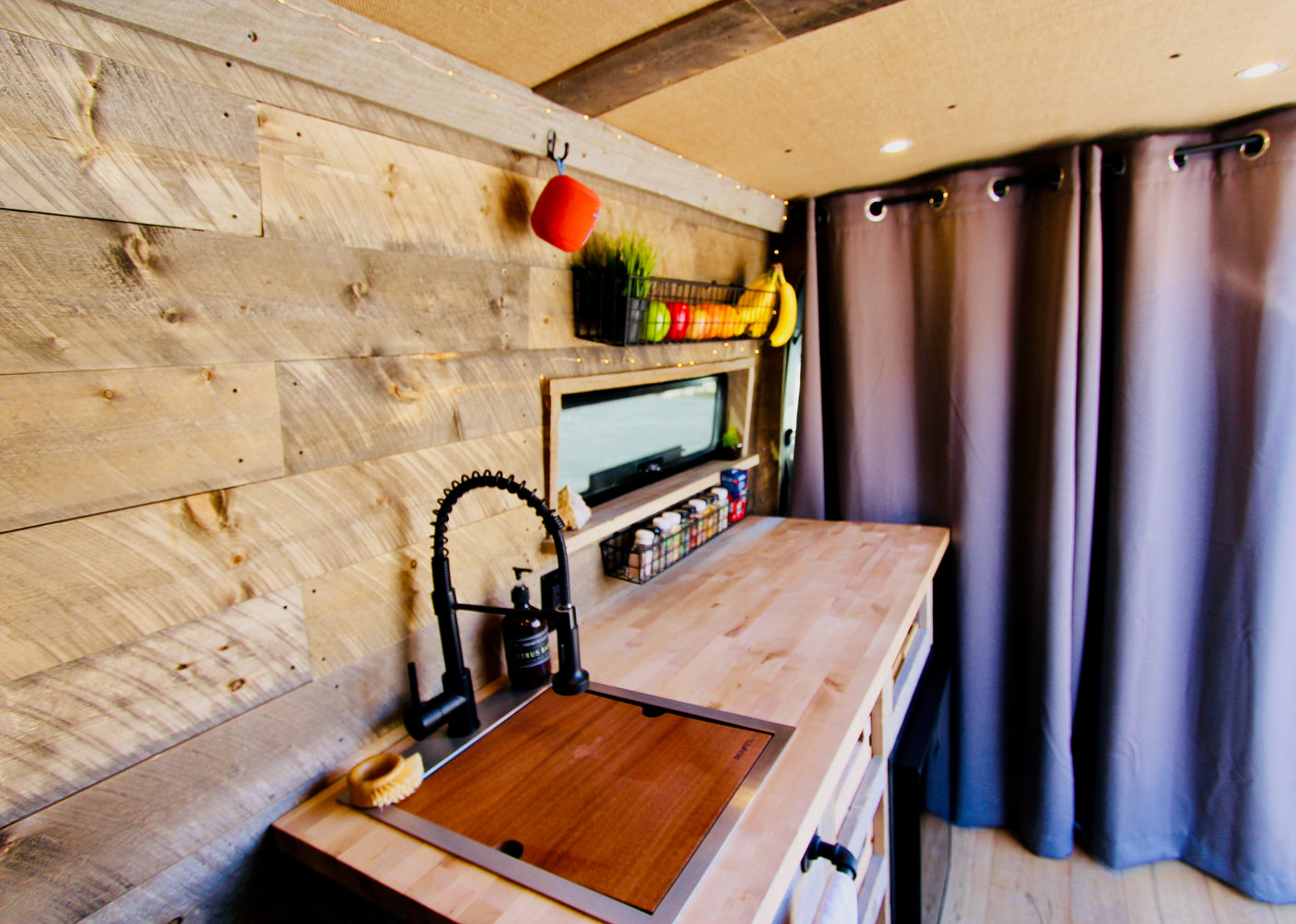 Cozy RV rental interior: wood-paneled kitchen with sink, countertop, spices, and fruit basket. Ideal for campervan living.