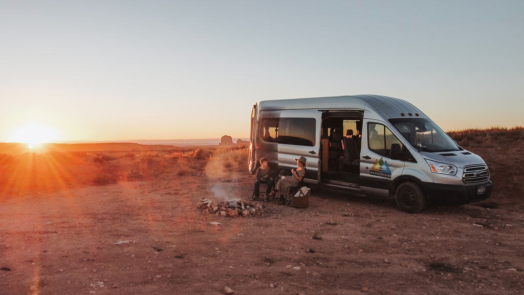 parked in a desert-like setting with adults by the van and kids at a campfire.|Silver RV rental (Wandervans camper van) at sunset. Two people sit by a campfire in the desert landscape