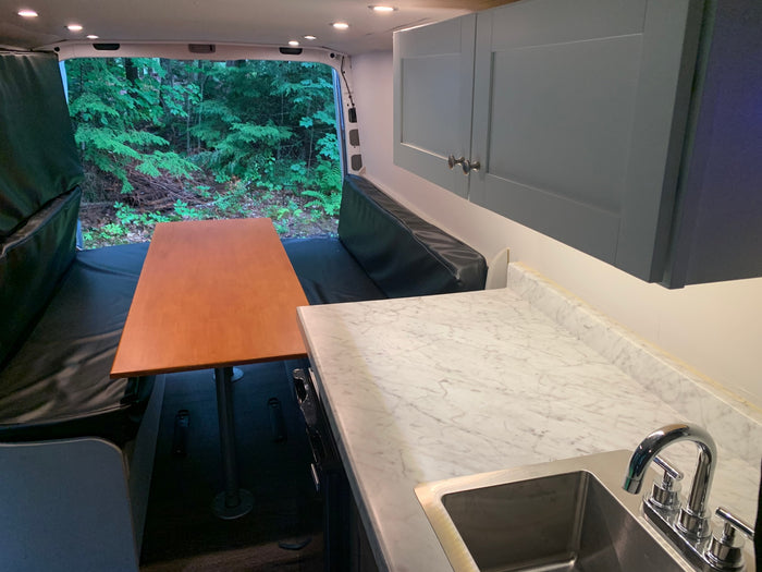 RV rental interior featuring a kitchen with sink, dining area with table & benches, and a forest view from the back.