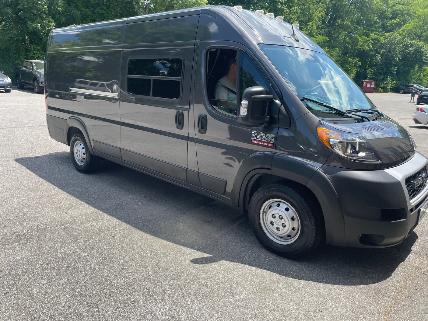 Dark gray Ram ProMaster camper van for RV rental, parked on asphalt with trees in background.