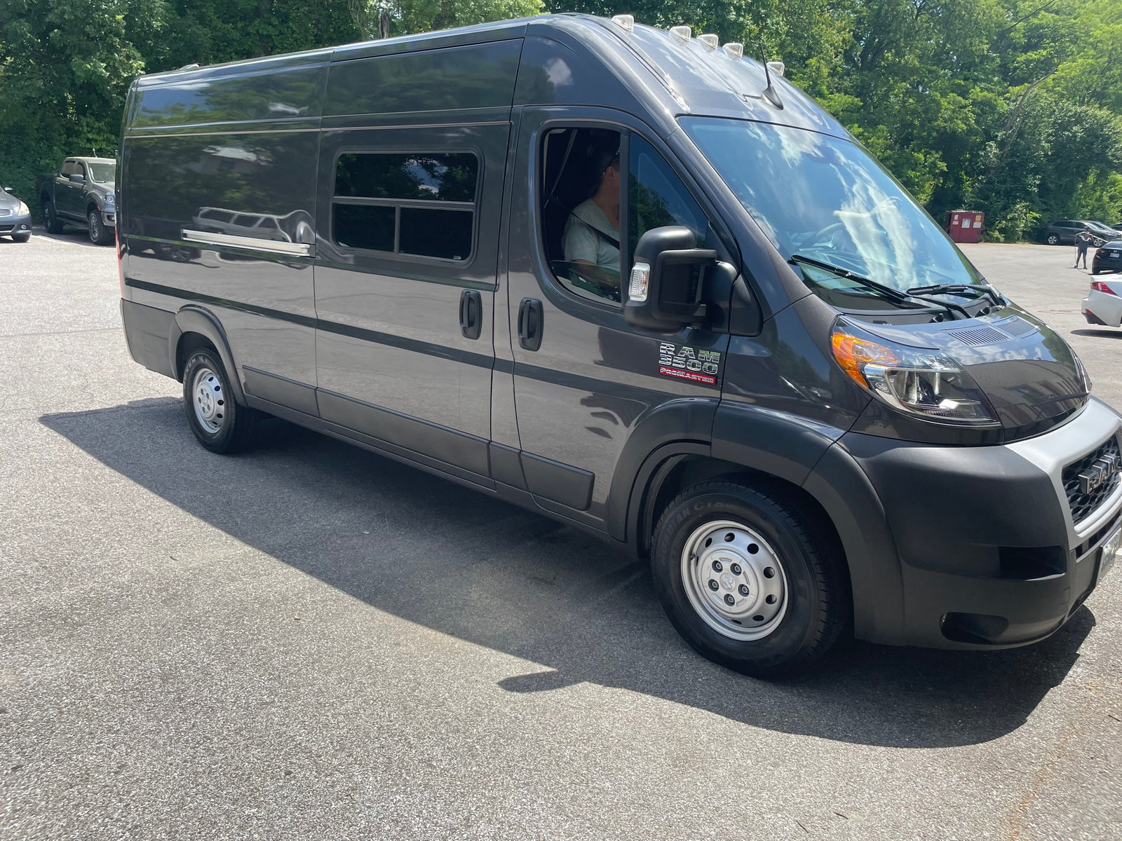 Dark gray Ram ProMaster camper van for RV rental, parked on asphalt with trees in background.