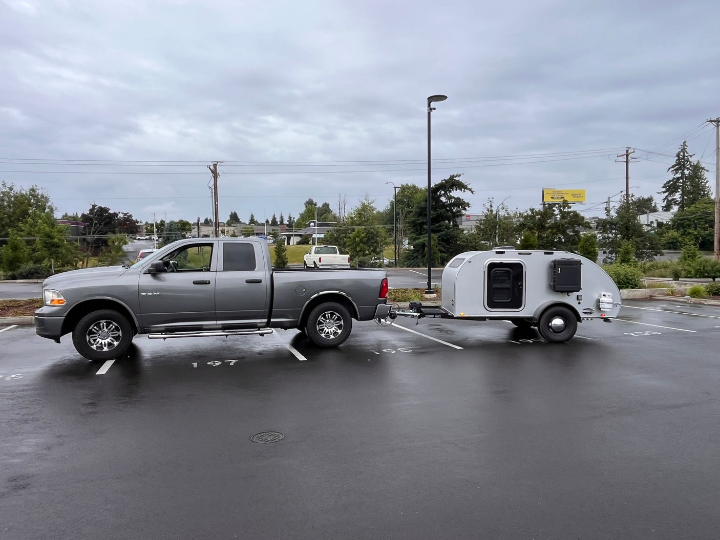 A gray pickup truck tows a compact silver teardrop RV trailer in a wet parking lot