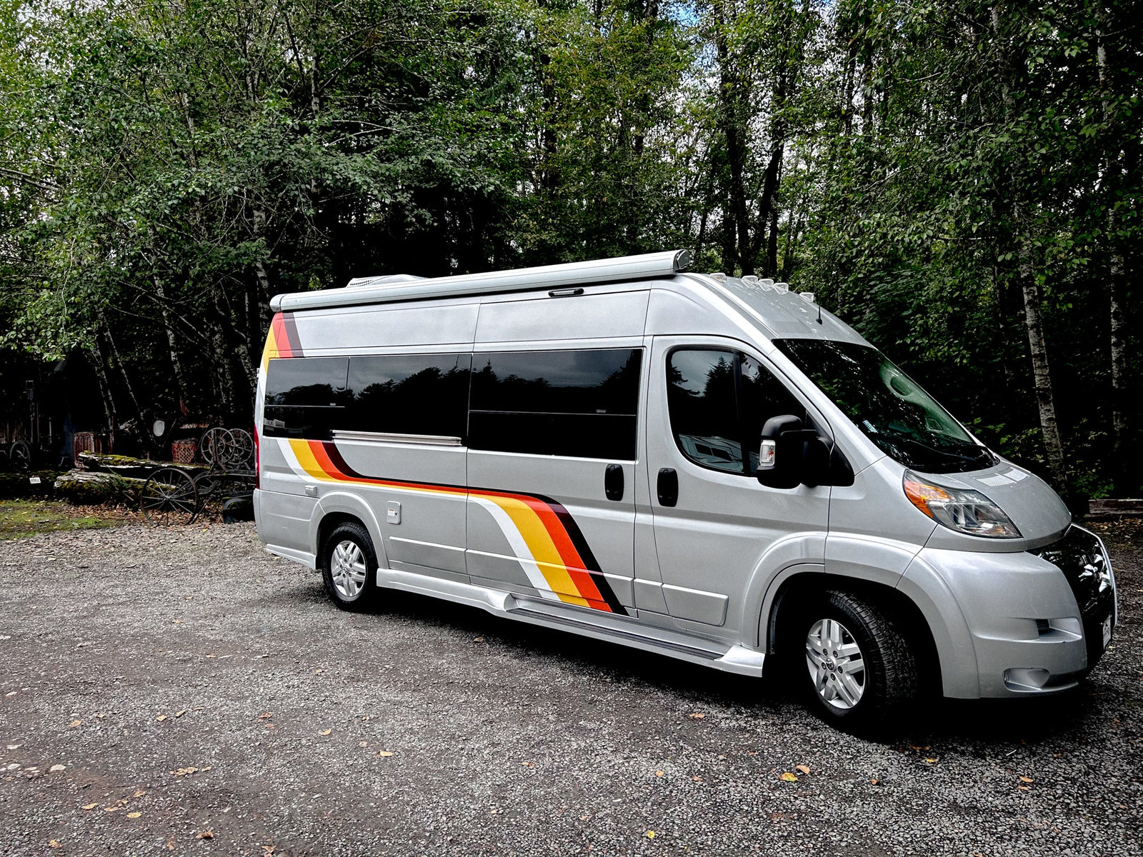Silver RV rental with colorful stripes parked on a gravel lot amidst green trees. This camper van is ready for your adventure.