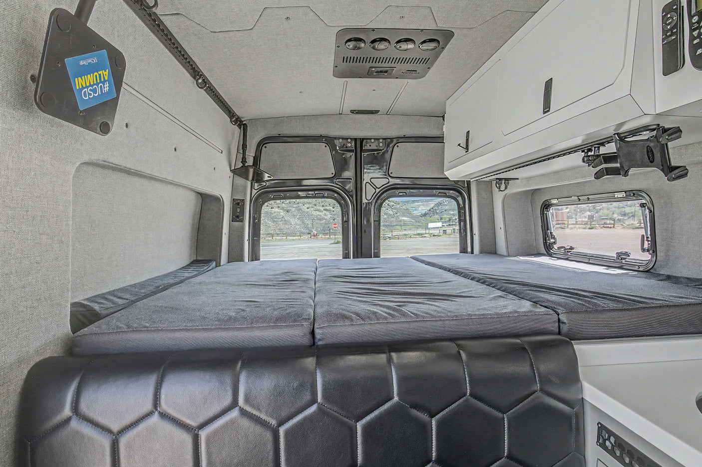 Spacious rv rental van interior: back view of the large bed, black leather bench, white cabinets, & mountain road scenery.