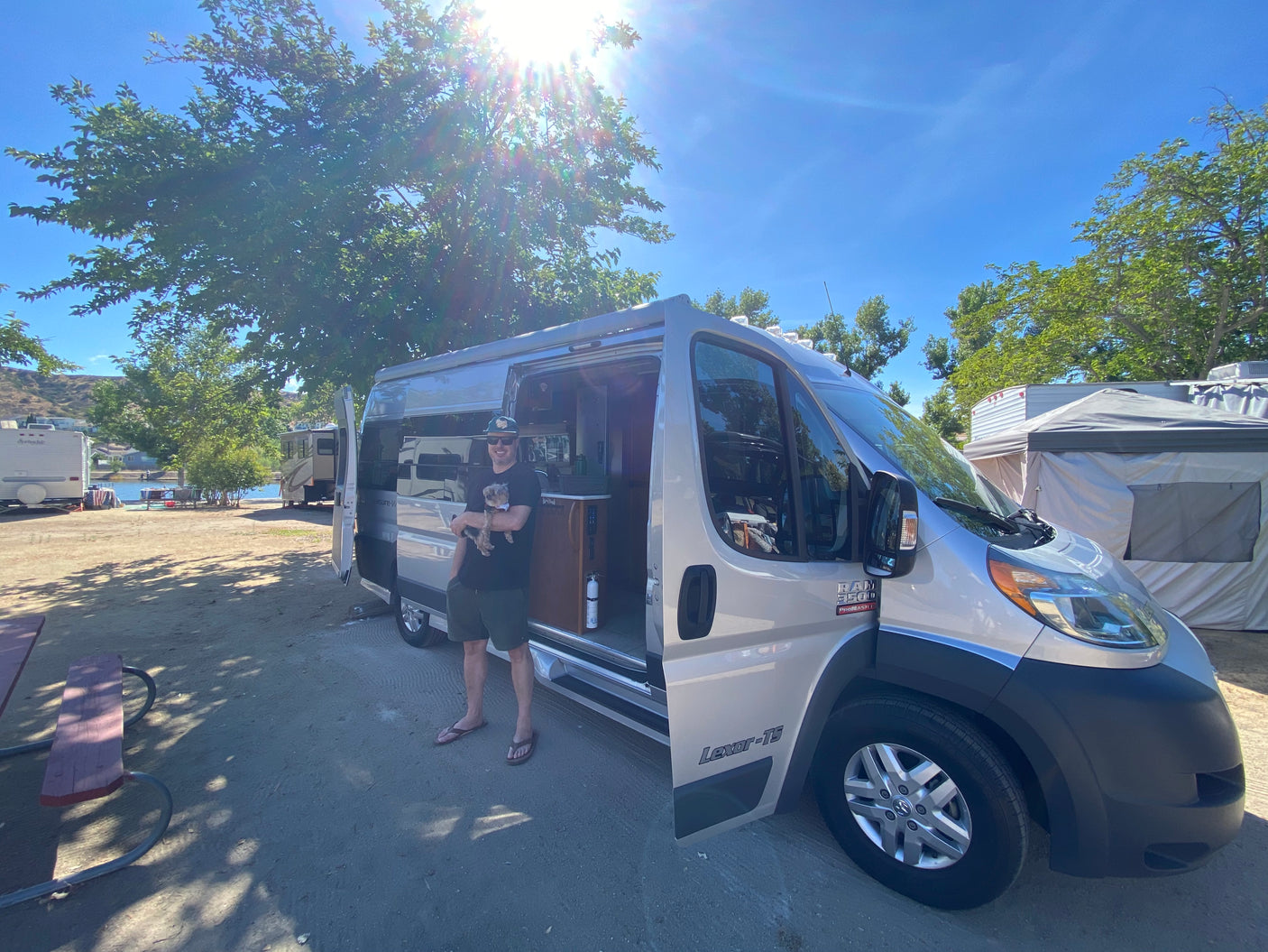 Man with dog by a silver RV rental at a sunny lakeside campsite, perfect for a summer road trip.