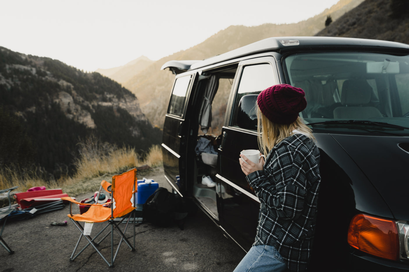 Scenic mountain camping with an RV rental. A woman sips coffee beside her black camper van and outdoor gear.