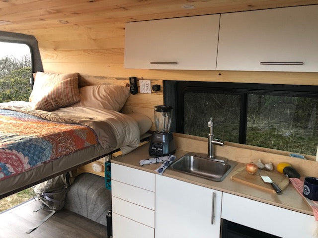 This RV rental's interior boasts a cozy elevated bed & functional kitchen with a sink, perfect for compact van life adventures.