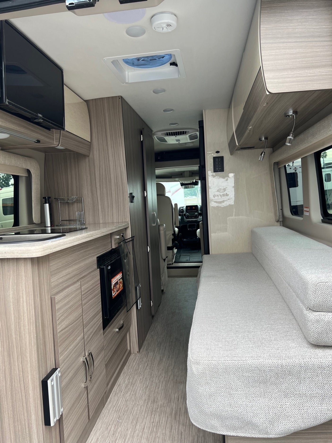 RV rental interior: Modern camper van layout with light wood cabinetry, kitchenette, mounted TV, and a comfy light grey sofa.