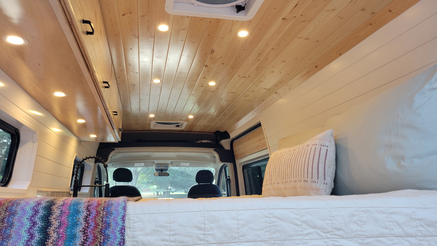 Inside an RV rental: cozy bed with colorful blanket, wooden ceiling lights, modern kitchen faucet, and front cabin view.