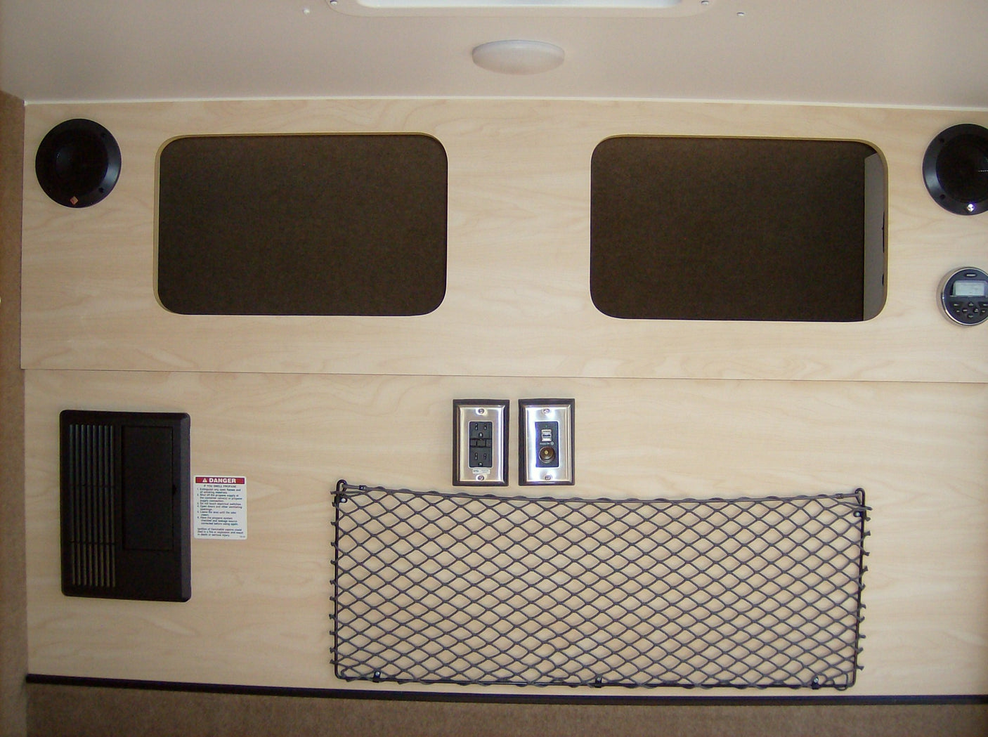 This RV rental interior shows light wood panels, overhead storage, speakers, power outlets, and a propane safety warning.