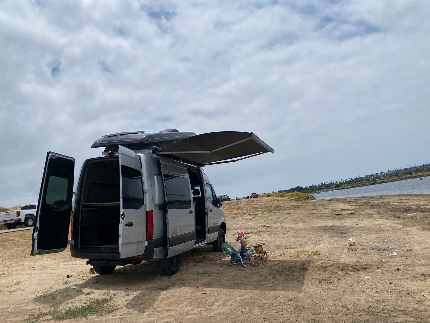 Silver RV rental van parked by the water, doors open, awning extended. A child relaxes in a chair on the sandy ground under a cloudy sky.