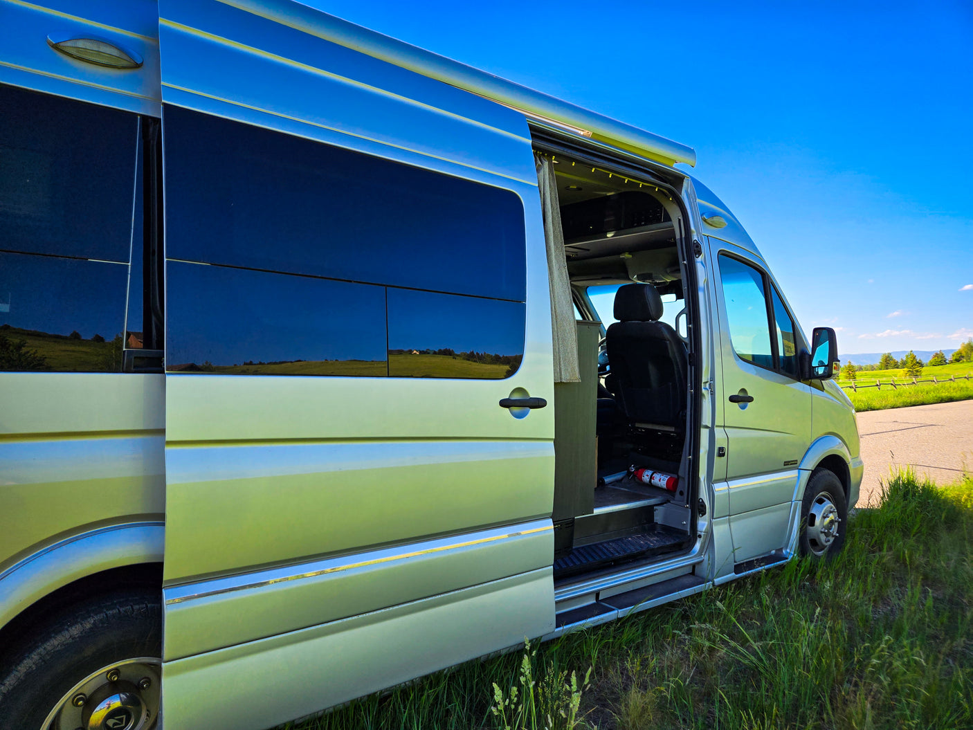 Silver RV rental van parked with its door open, showing interior and reflecting a scenic outdoor landscape.