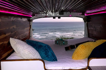 Cozy RV rental van interior with bed facing ocean view. Wood-paneled walls, pink LED lights, and a ukulele create a relaxed travel vibe.