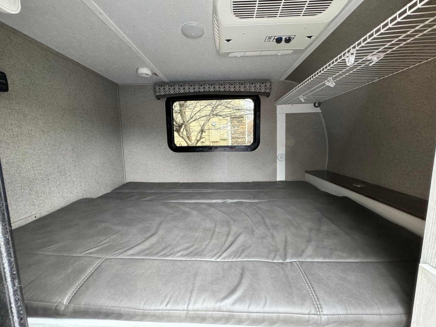 Interior of an RV rental showing a comfy bed, window, overhead AC, and storage for mobile living.