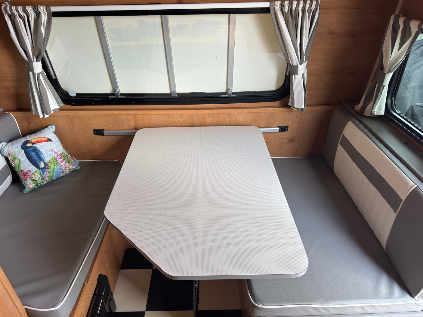 This cozy rv rental features a bright dining area with a light patterned table, gray benches, striped curtains, and a vibrant toucan pillow.