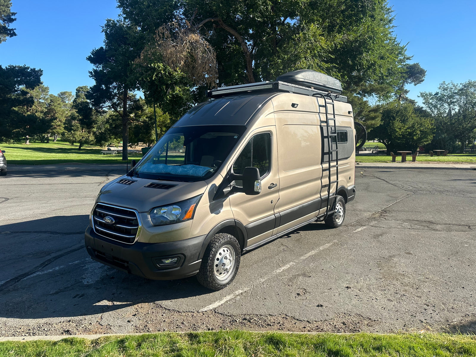 Adventure-ready tan Ford Transit camper van RV rental, equipped with a roof rack and ladder, parked in a sunny park.