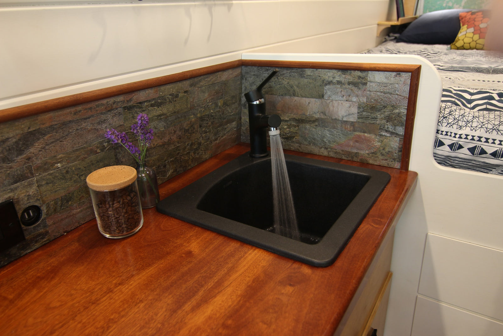 Modern RV rental kitchen with a black sink, running water, and stone backsplash. Features a wooden counter, coffee beans, and lavender for cozy living.