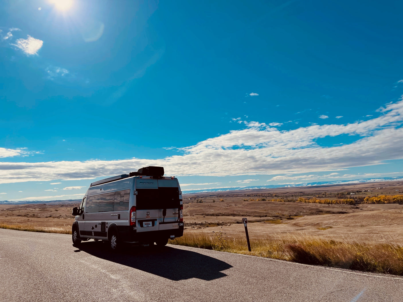 A modern RV rental parked on a scenic road, overlooking golden plains and distant snow-capped mountains under a bright sky.