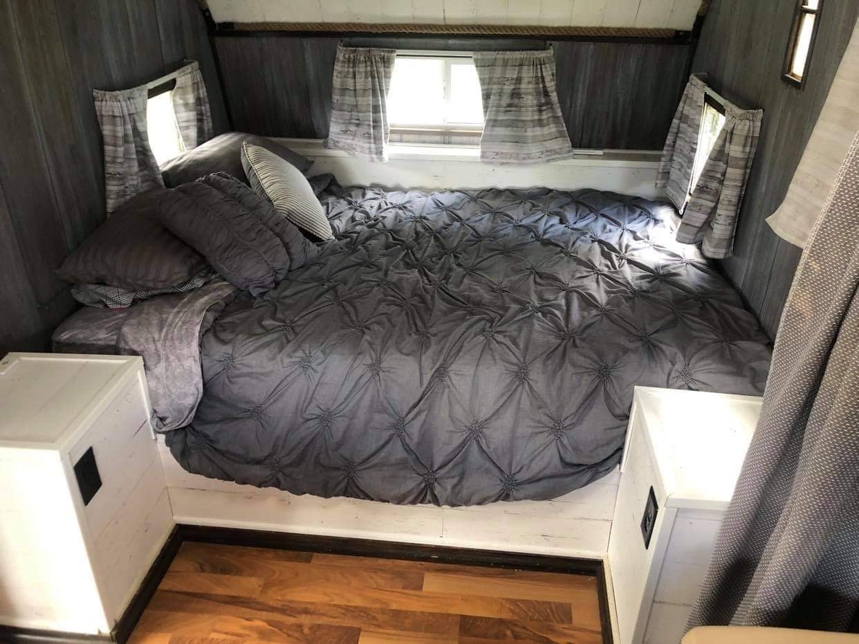 An inviting RV rental bedroom. Features a spacious gray tufted bed, multiple pillows, and side windows with curtains.