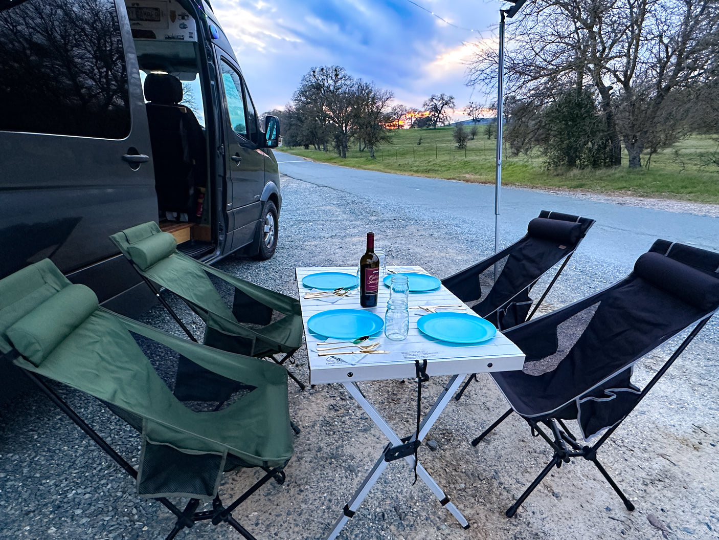 Scenic RV rental experience: Portable dining table with blue plates, wine, and camping chairs set beside a camper van at sunset.