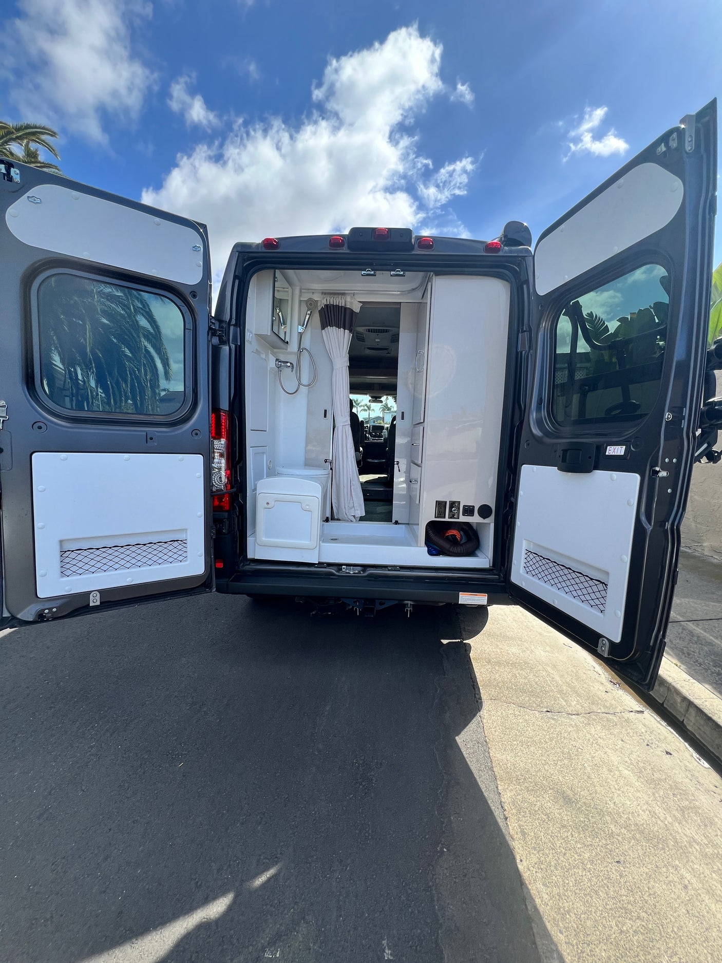 Open rear doors of a dark grey RV rental reveal a compact interior with a shower, toilet, and storage. Parked on asphalt.
