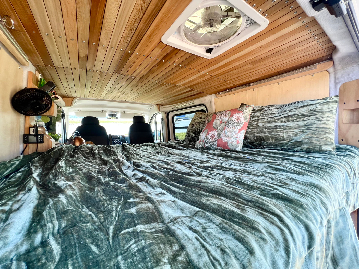 RV rental camper van interior: A cozy bed with green crushed velvet bedding, wooden slatted ceiling, and roof vent fan.