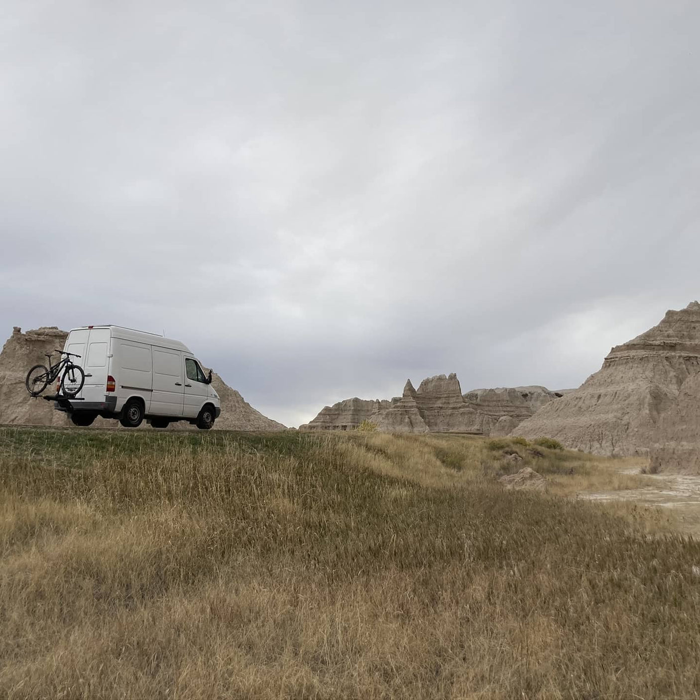 White Sprinter van with bike rack parked in front of striking badlands formations. Ideal for an RV rental road trip!
