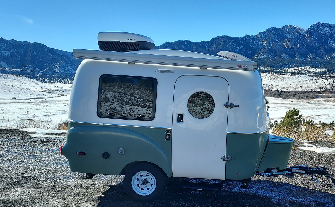 An RV rental: a white & green camper with Dometic AC & Fiamma awning, parked near snowy mountains under a clear sky.