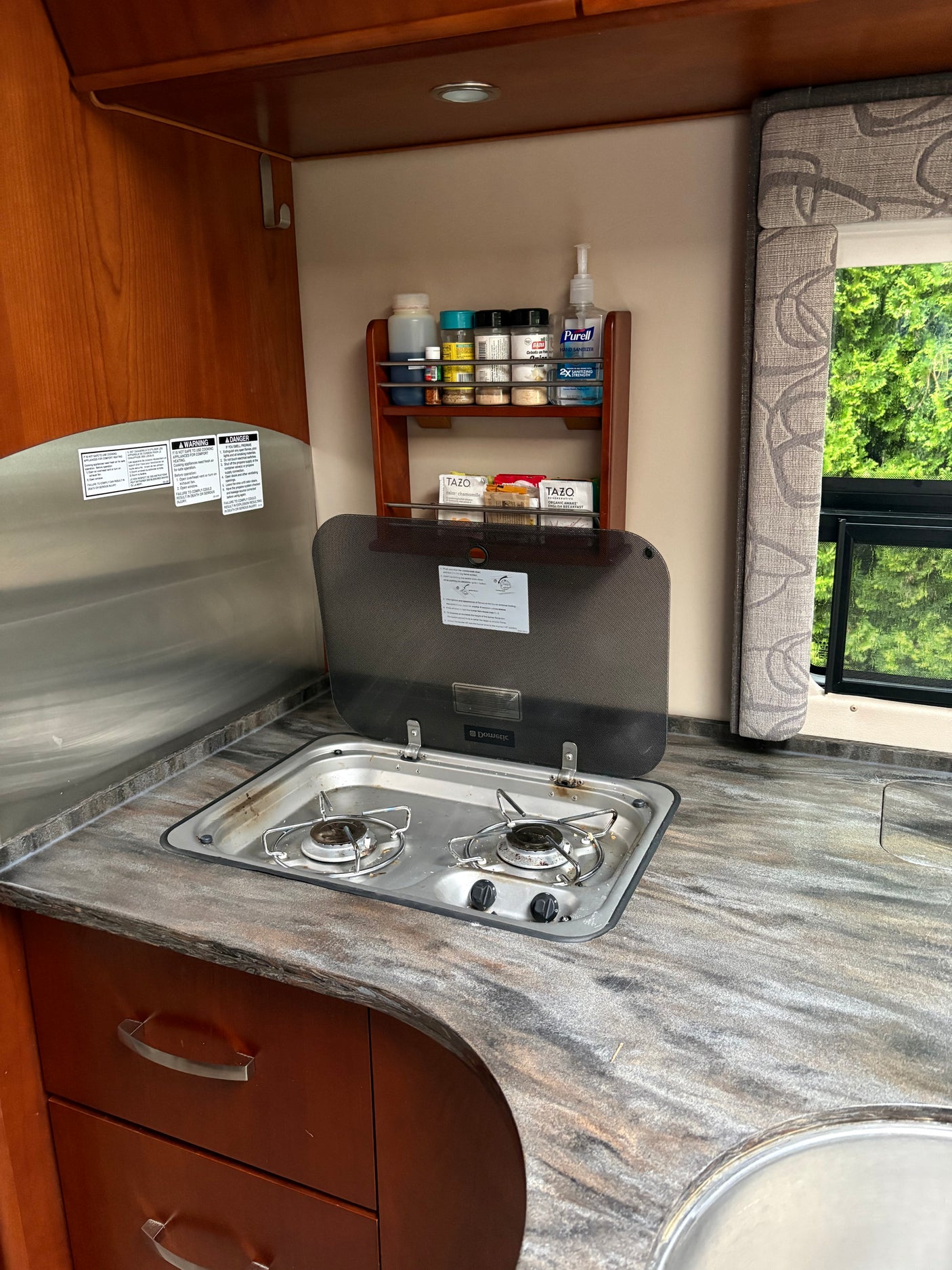 RV rental kitchen featuring a two-burner stove, granite-look counter, a shelf with spices & Tazo tea, and a window with nature views.