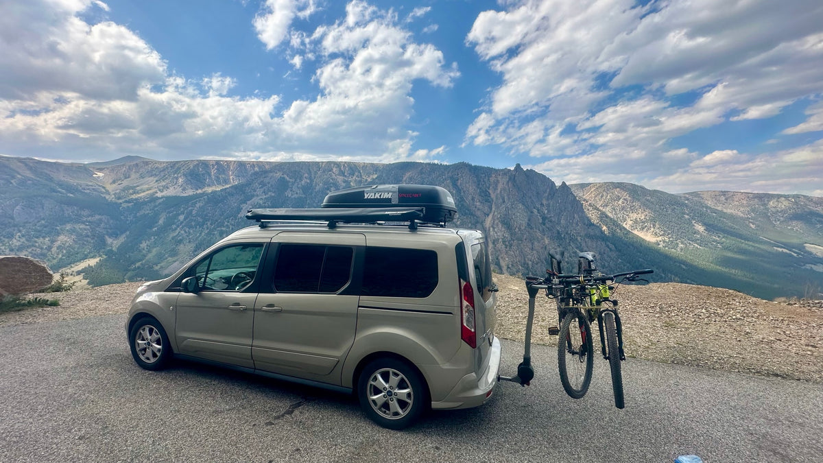 This RV rental van, loaded with bikes, roof cargo box, and awning, sits on a mountain road under a cloudy sky, ready for adventure. — photo 14