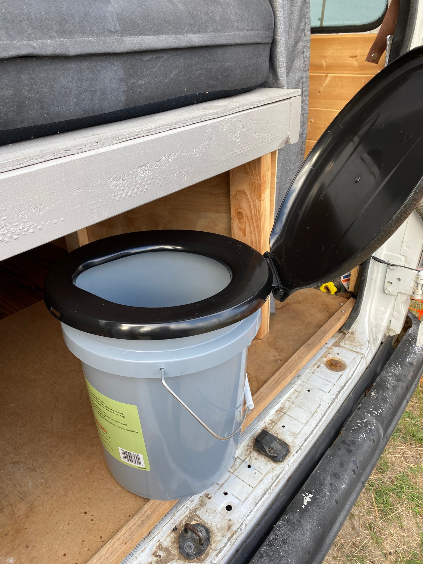 A gray portable bucket toilet with a black seat and lid is set up inside an RV rental, ready for travel convenience.