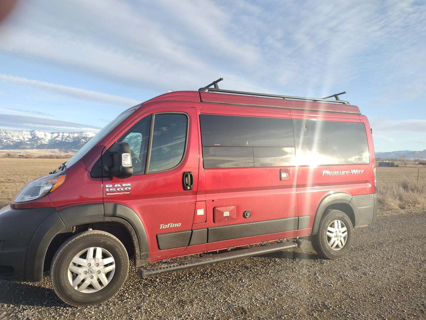 Red Pleasure-Way Tofino camper van, an ideal RV rental, parked on a gravel road with snowy mountains in the background.