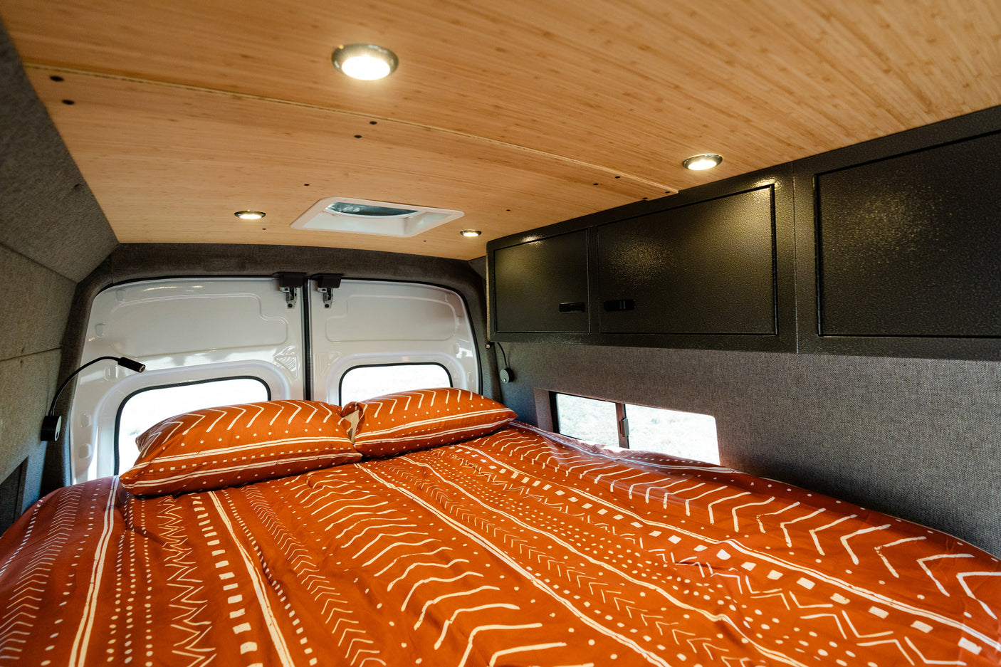 Comfortable RV rental bed with patterned bedding, wood ceiling, overhead cabinets, and functional lighting.