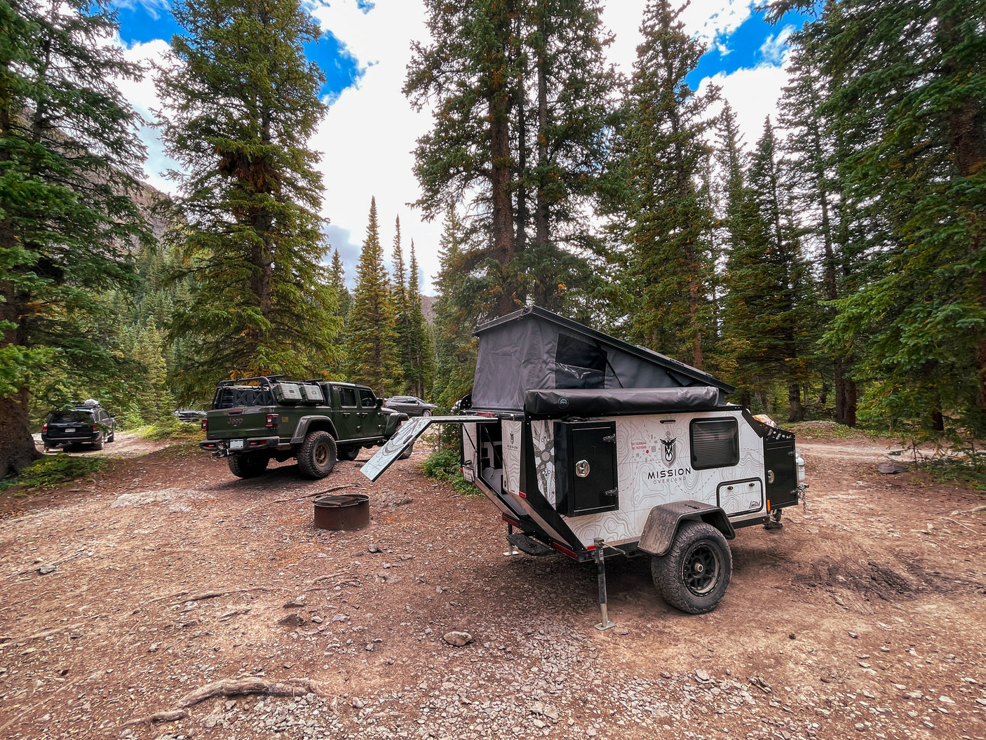 A pop-top Mission Overland trailer and Jeep Gladiator are set up for an outdoor RV rental adventure in a forest.