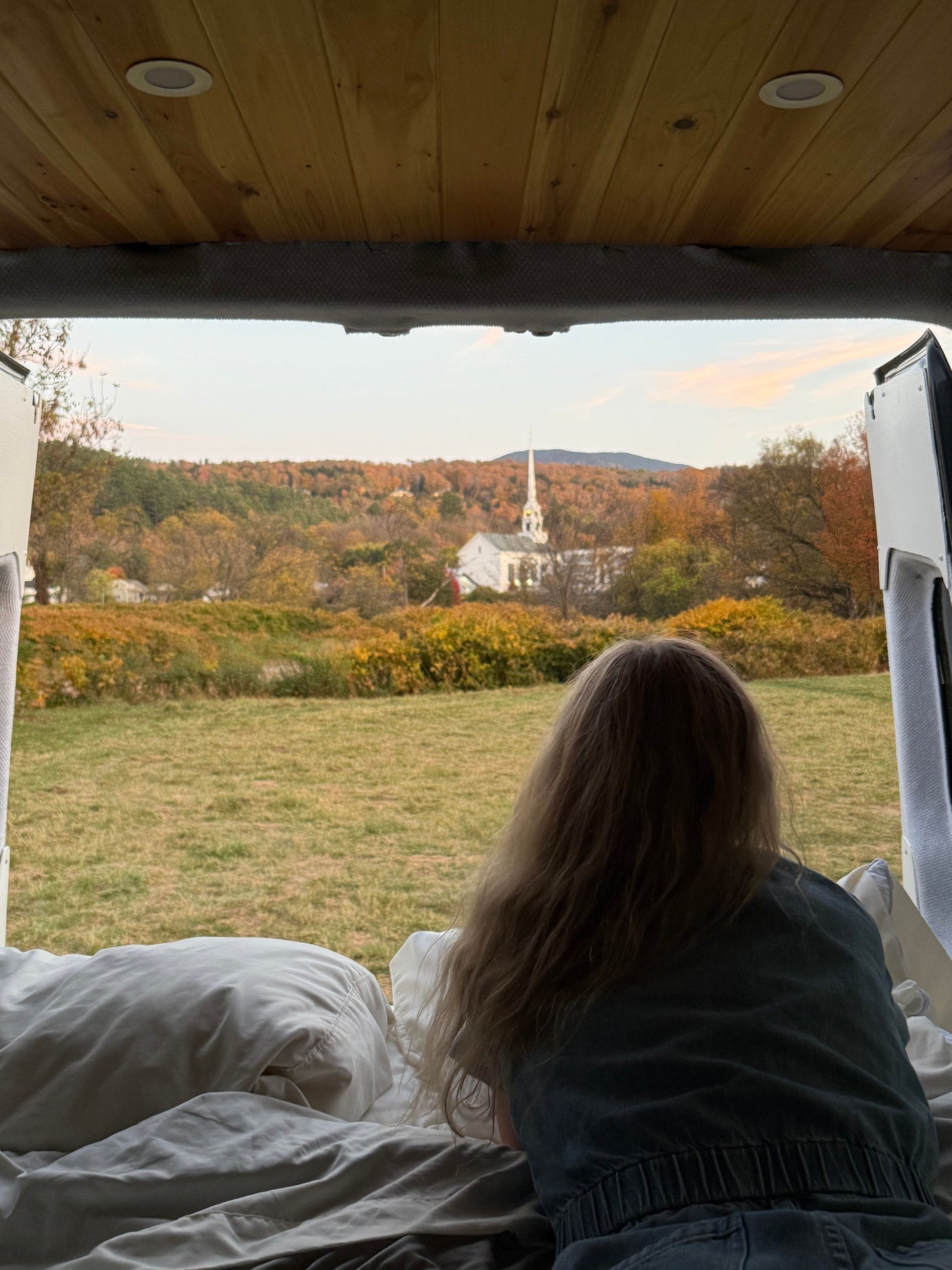 Cozy rv rental view: A person relaxes on a bed, looking out at an autumn landscape with a white church & vibrant fall foliage.