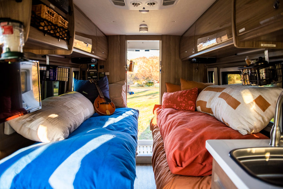 Bright RV rental interior: two cozy beds, one with a mandolin, overhead storage, and a sink, overlooking a scenic outdoor view.