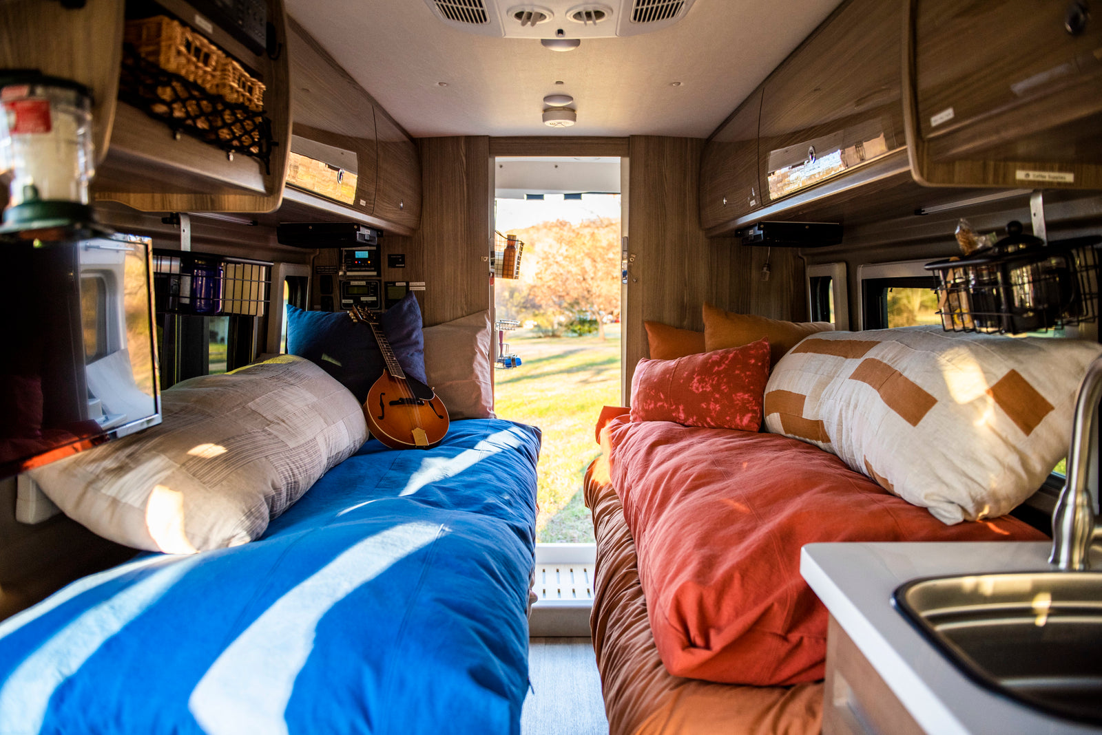 Bright RV rental interior: two cozy beds, one with a mandolin, overhead storage, and a sink, overlooking a scenic outdoor view.