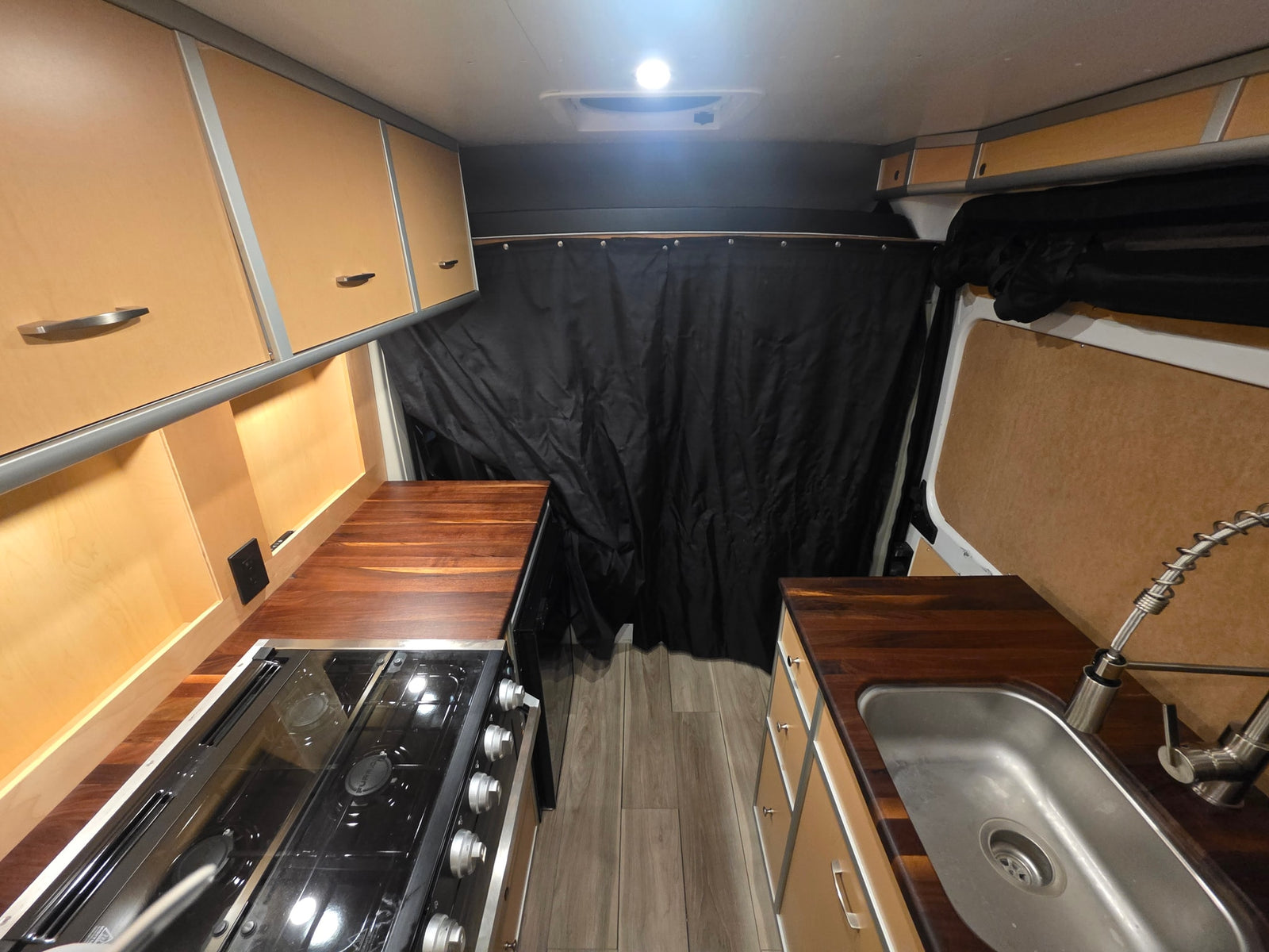 Modern RV rental kitchen with dark wood counters, stainless sink, stovetop, light cabinets, and black privacy curtains.