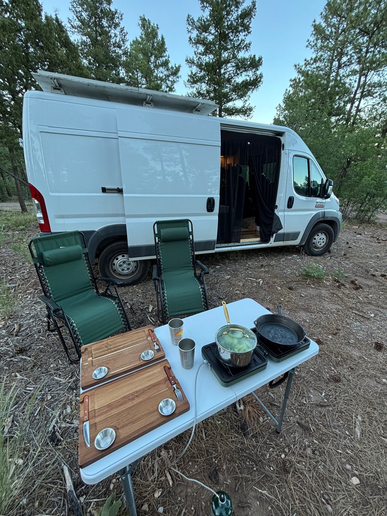 RV rental parked in a forest clearing with outdoor cooking setup: pot, pan, cutting boards, and chairs.