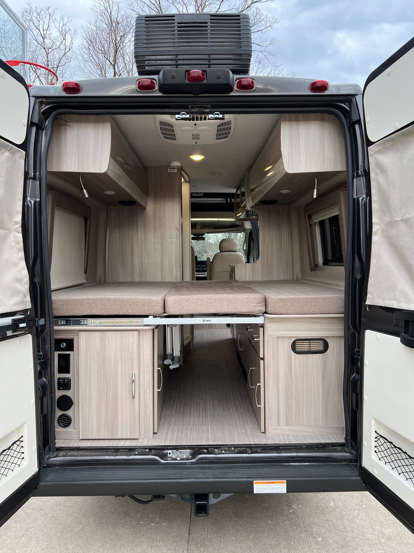 RV rental: View from open rear doors into a cozy camper van interior with modular bed, cabinetry, and flooring. Ready for adventure!