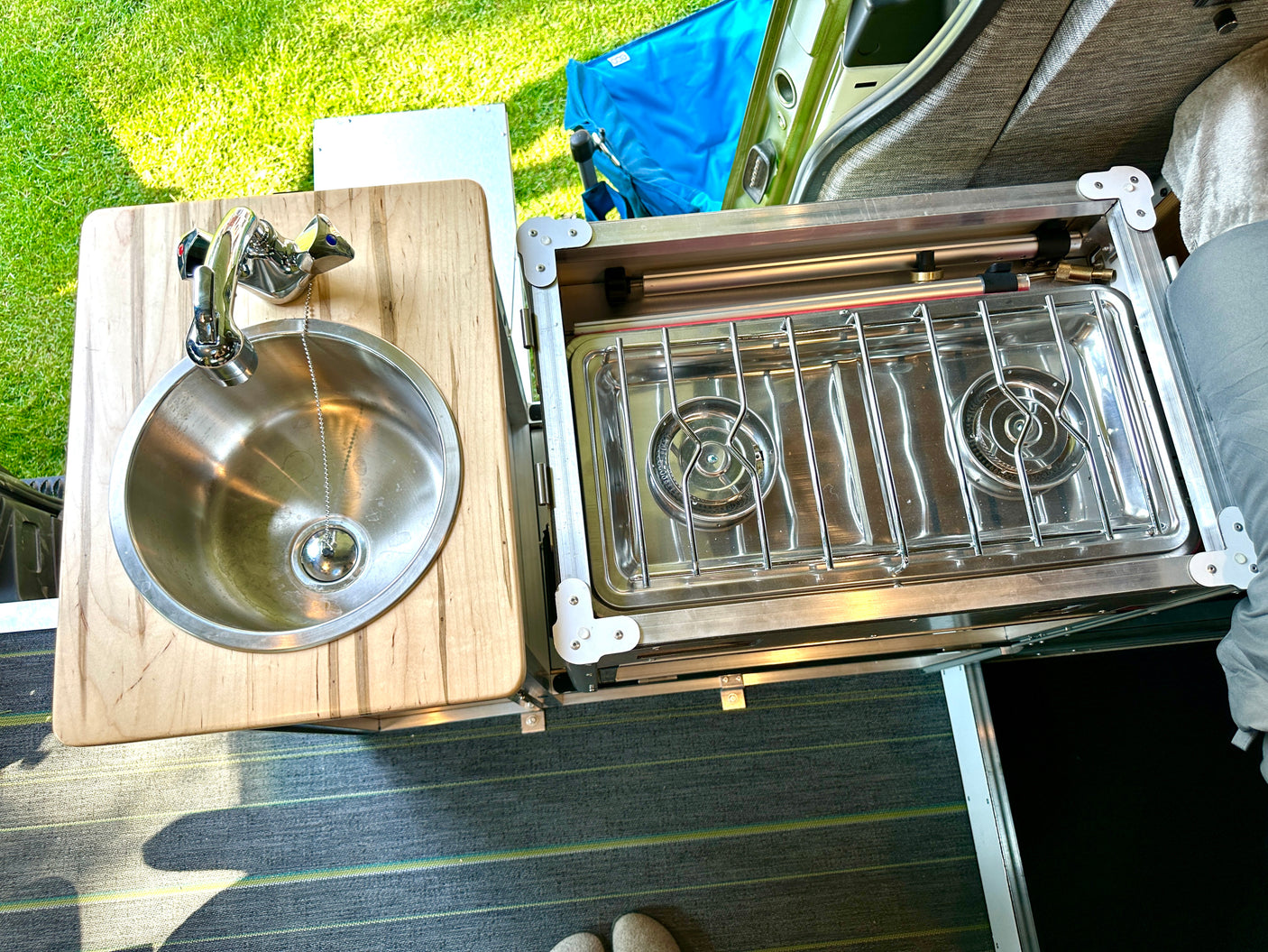 An rv rental's outdoor kitchen setup: wooden counter with sink and a two-burner stove, visible with green grass outside.