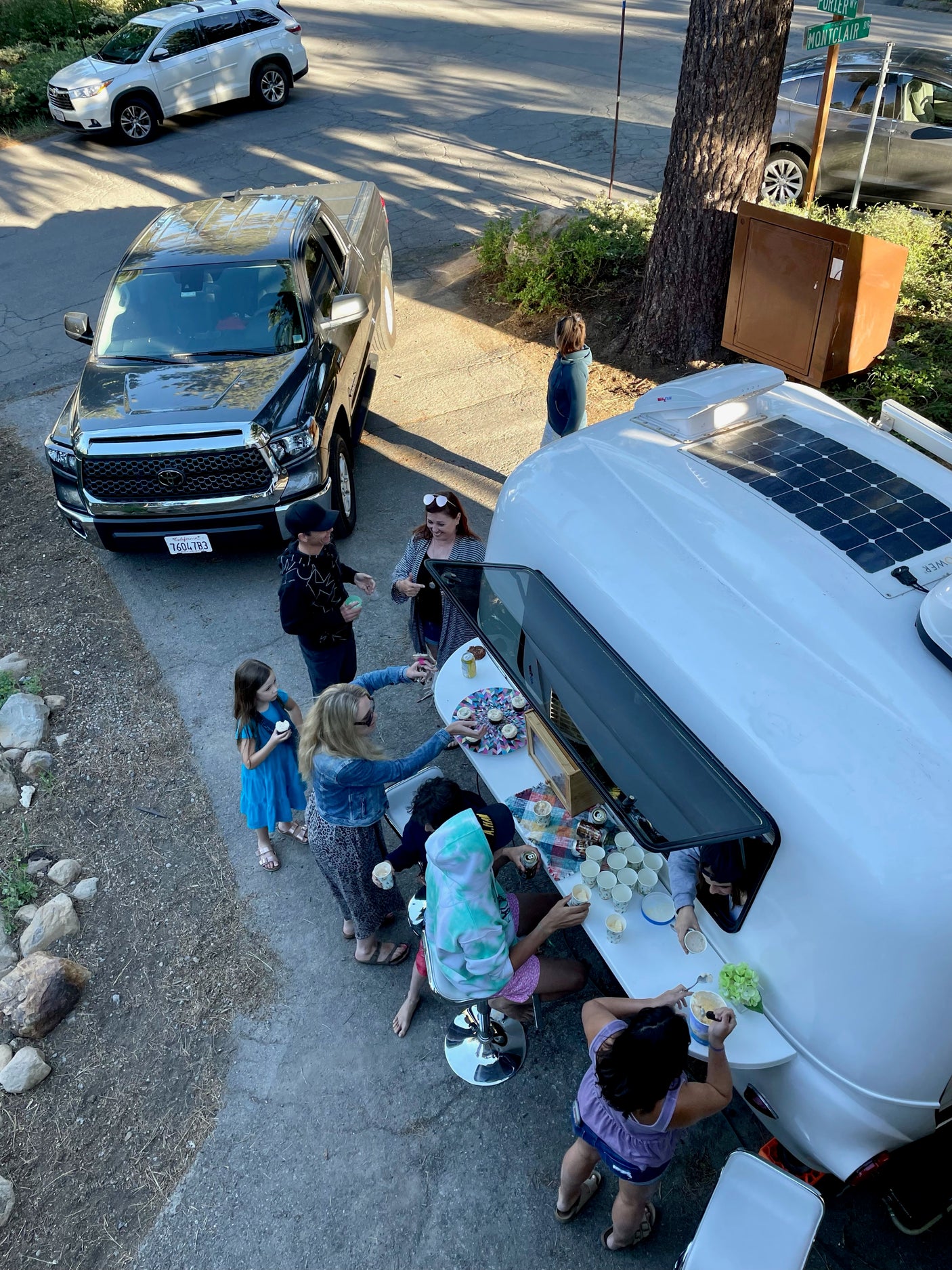Lively outdoor gathering enjoying ice cream and cupcakes served from a white RV rental's window, with cars parked nearby.