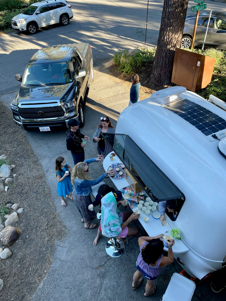 Lively outdoor gathering enjoying ice cream and cupcakes served from a white RV rental's window, with cars parked nearby.