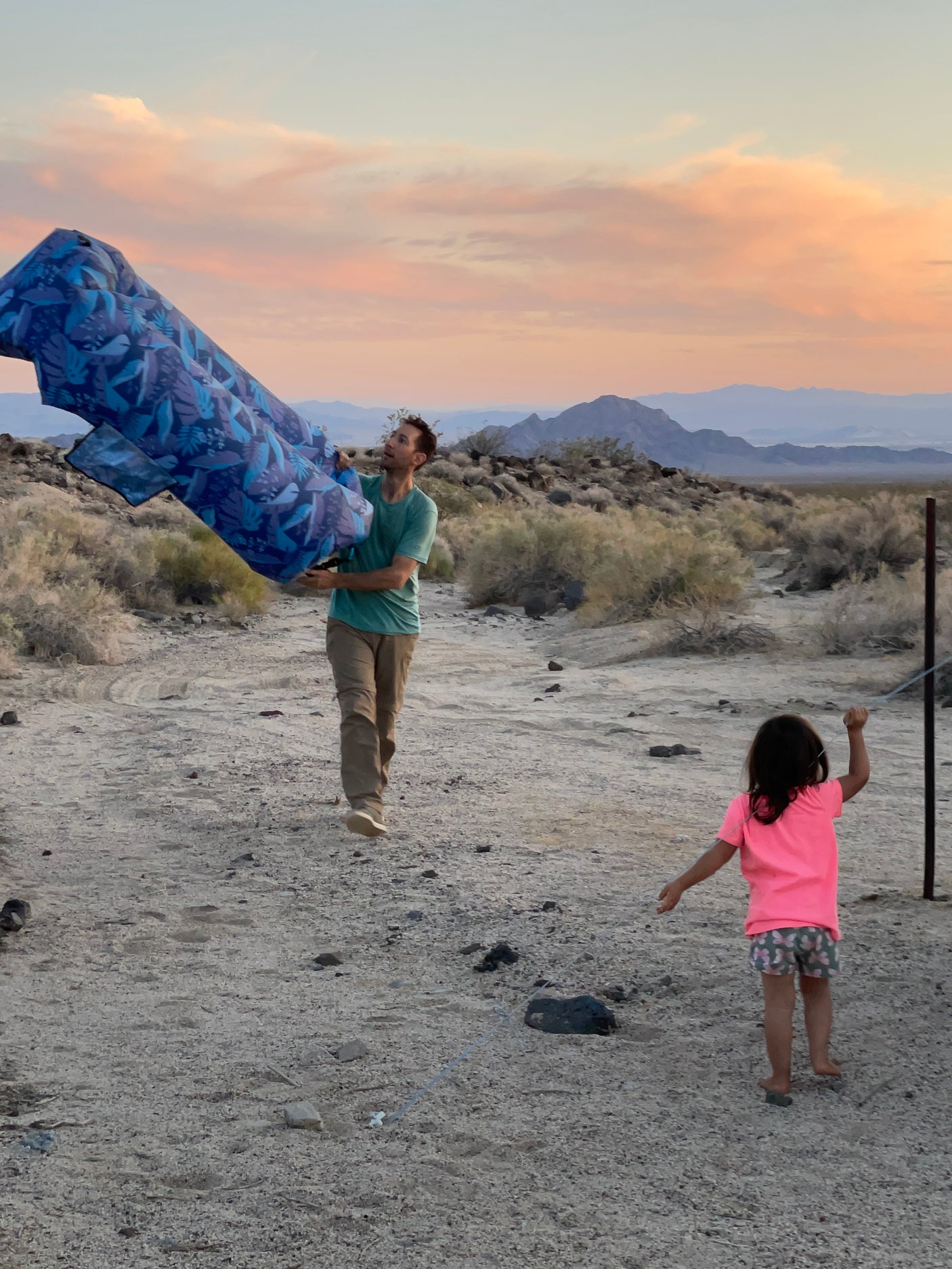 RV rental fun: A man and child enjoy a vast desert landscape and sunset, holding blue outdoor gear.