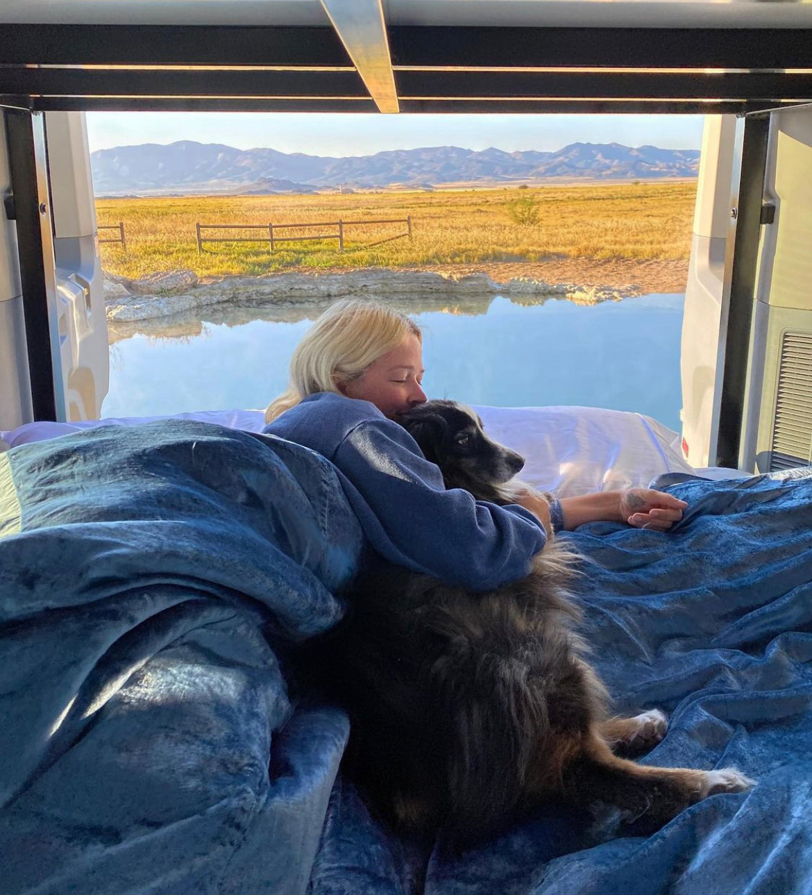 RV rental comfort: Woman & dog relaxing on bed in a van, with panoramic views of a tranquil lake, golden fields, and mountains.