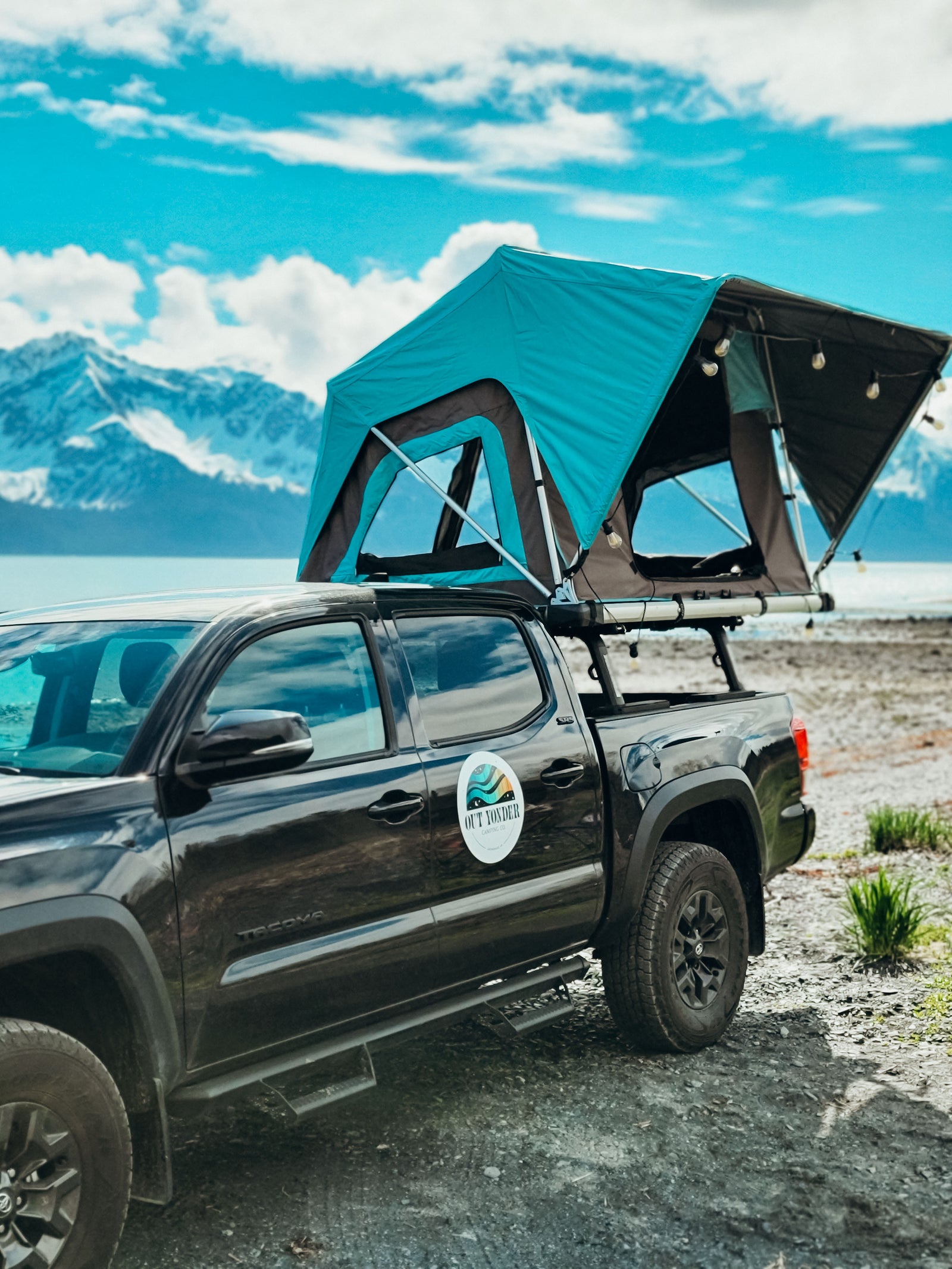 RV rental: Black pickup truck with a vibrant teal rooftop tent set up by a serene lake with majestic mountains beyond.