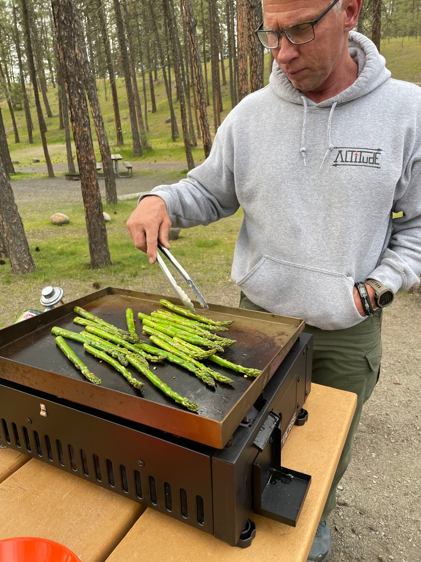 Man in grey hoodie cooking asparagus on a portable griddle at a scenic campsite. Ideal for your RV rental trip.