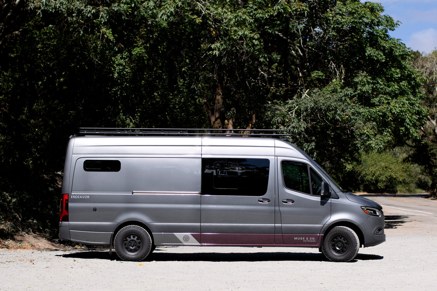 This grey custom Mercedes Sprinter van, an 'Endeavor' rv rental, is parked roadside surrounded by green trees.